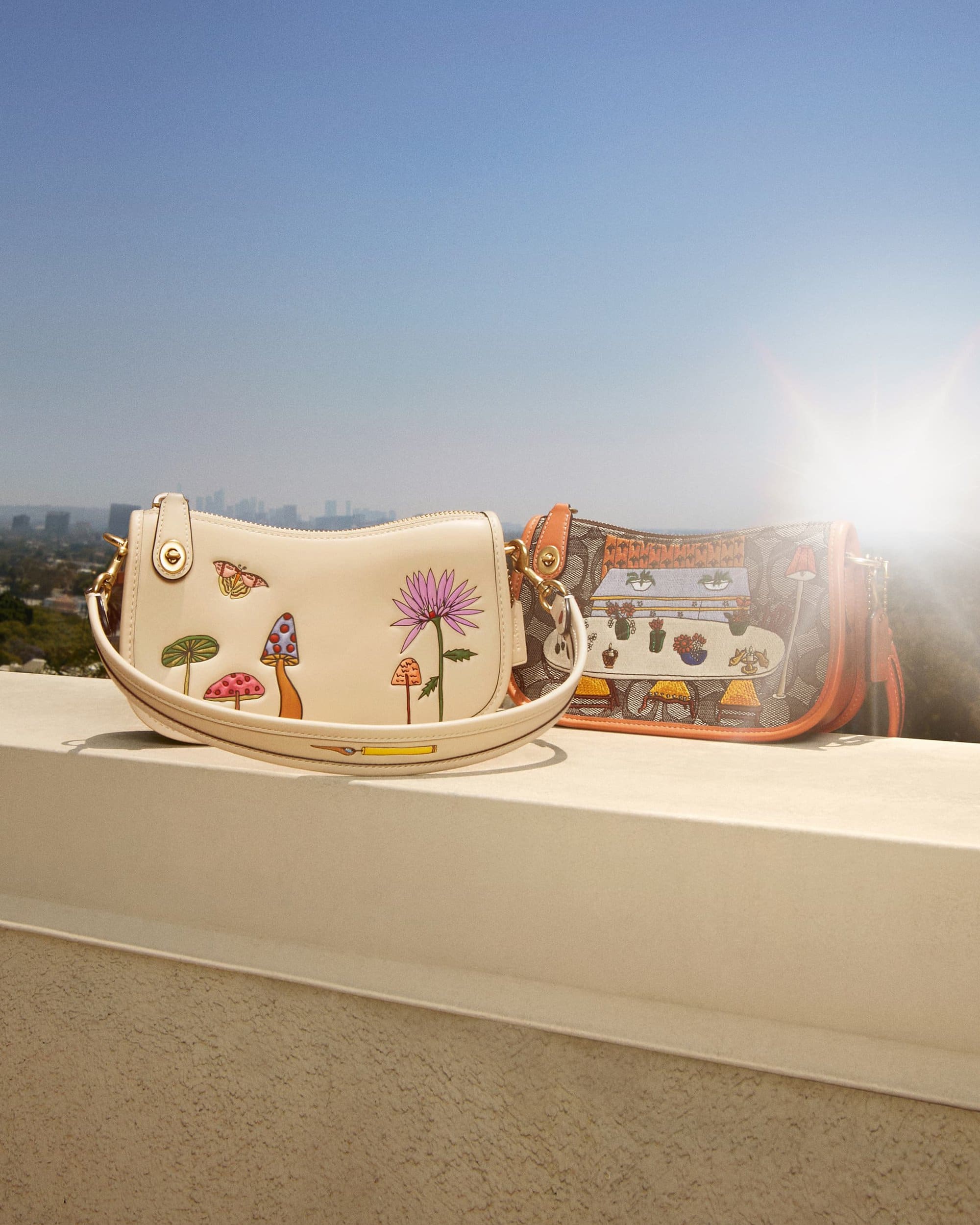 Two purses with floral and mushroom designs sit on a ledge with a cityscape and blue sky in the background on a sunny day.