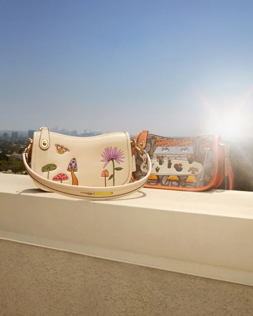 Two purses with floral and mushroom designs sit on a ledge with a cityscape and blue sky in the background on a sunny day.