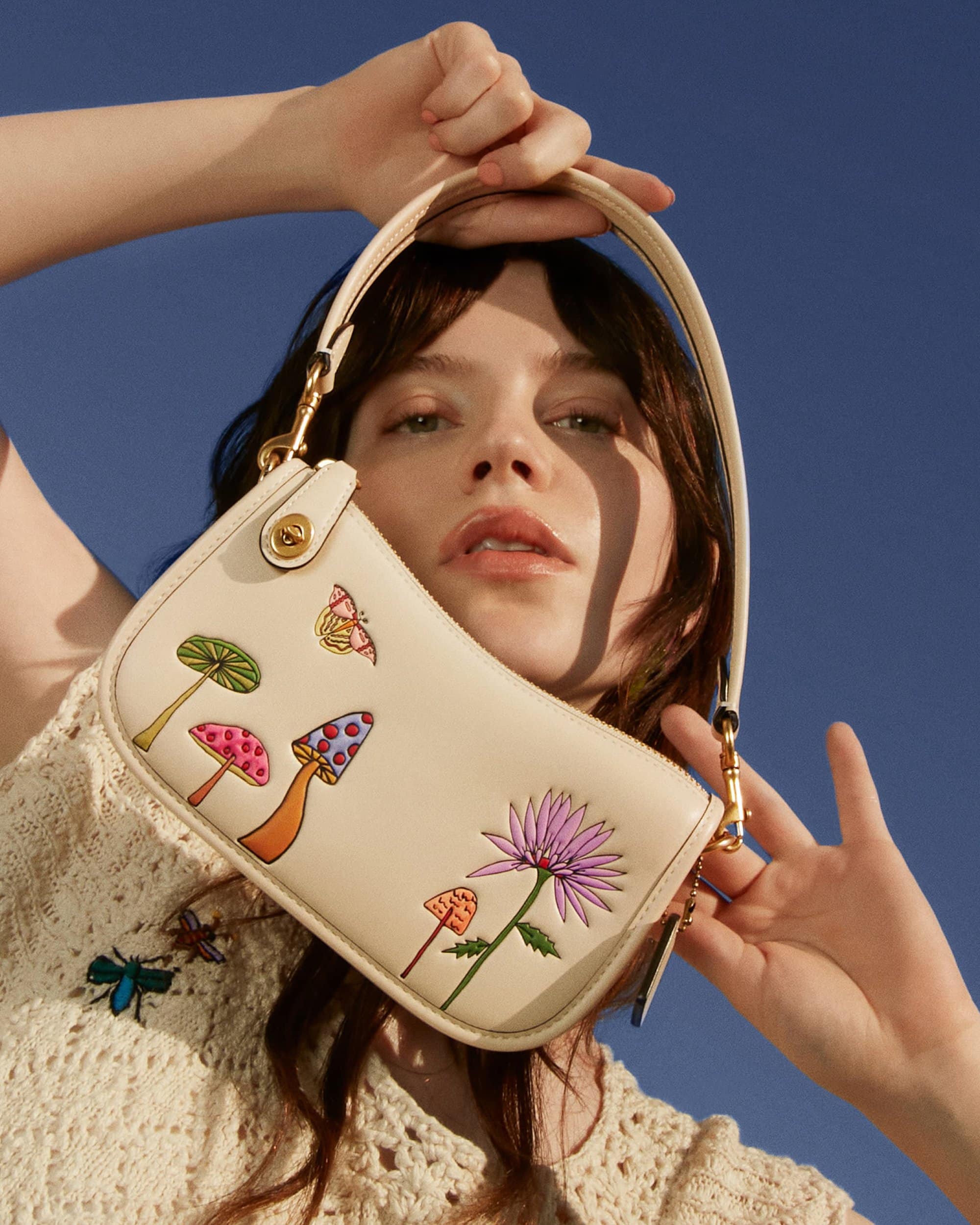 Woman holding cream-colored purse with mushroom and flower illustrations in front of a clear blue sky in a fashion setting.