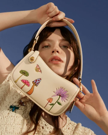 Woman holding cream-colored purse with mushroom and flower illustrations in front of a clear blue sky in a fashion setting.