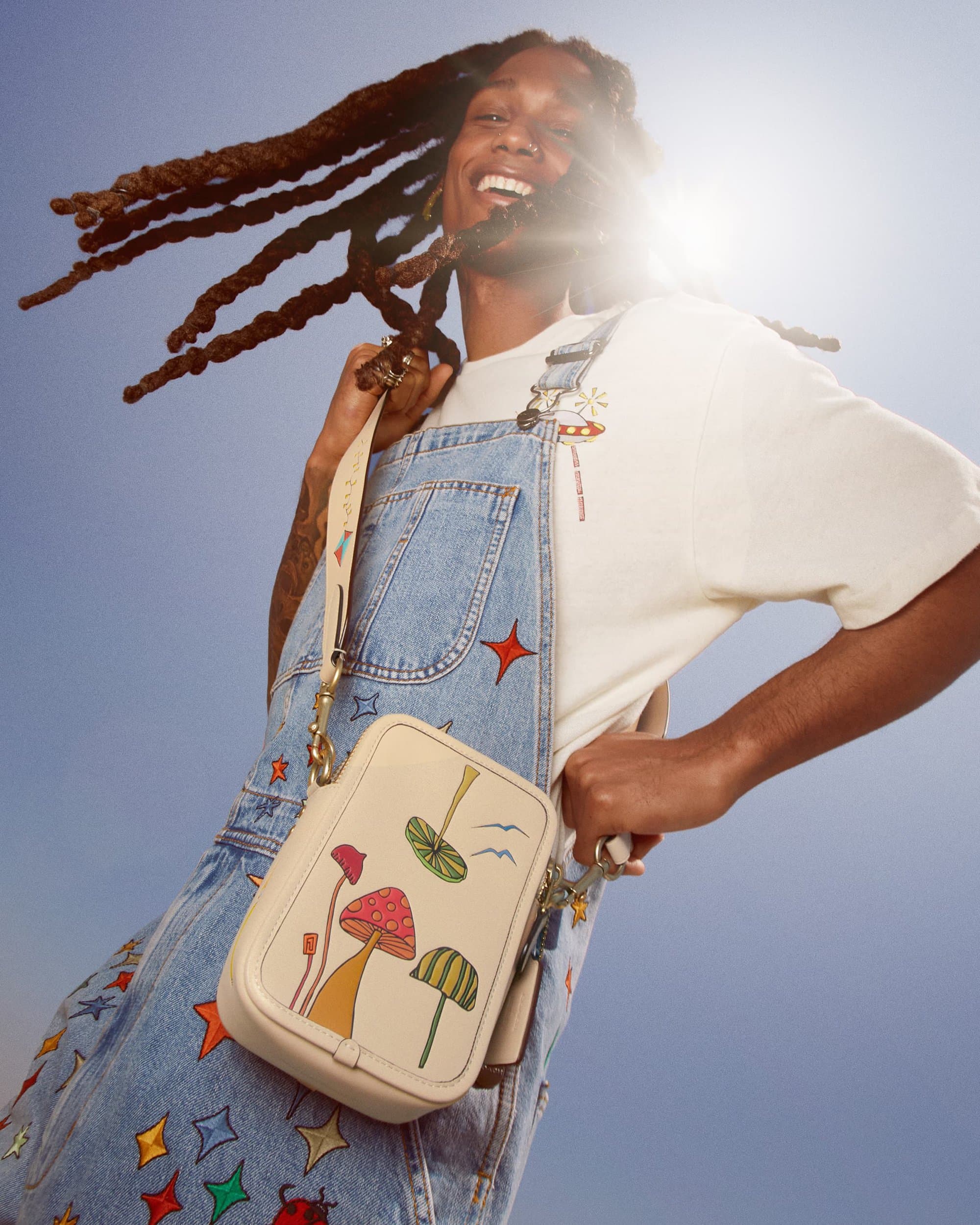 A joyful adult with dreadlocks wears denim overalls with an illustrated bag, posing in front of a bright sky with a radiant smile.