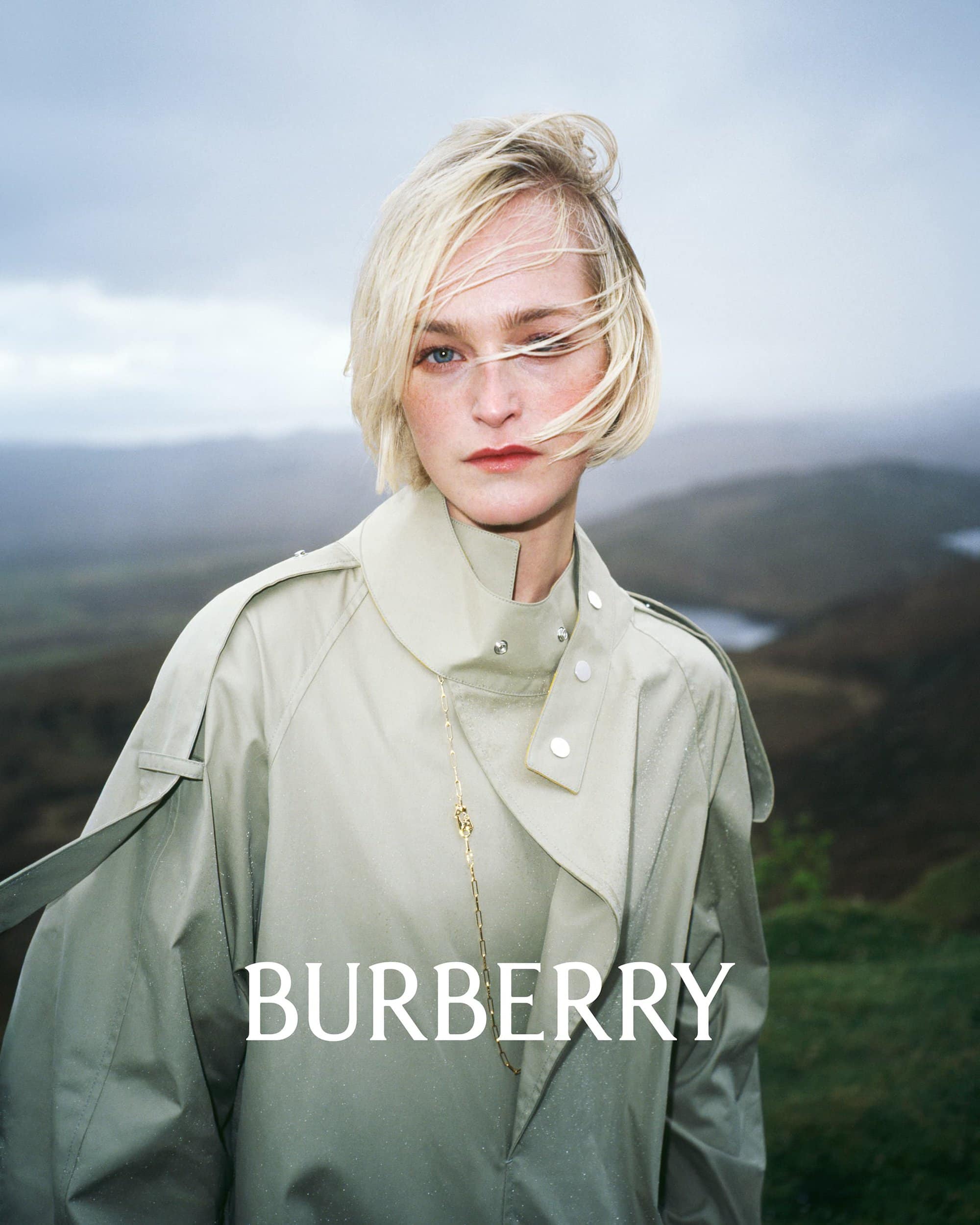 Fashion shot of a blonde woman in a light trench coat standing outdoors, landscape in the background. Burberry logo in the foreground.