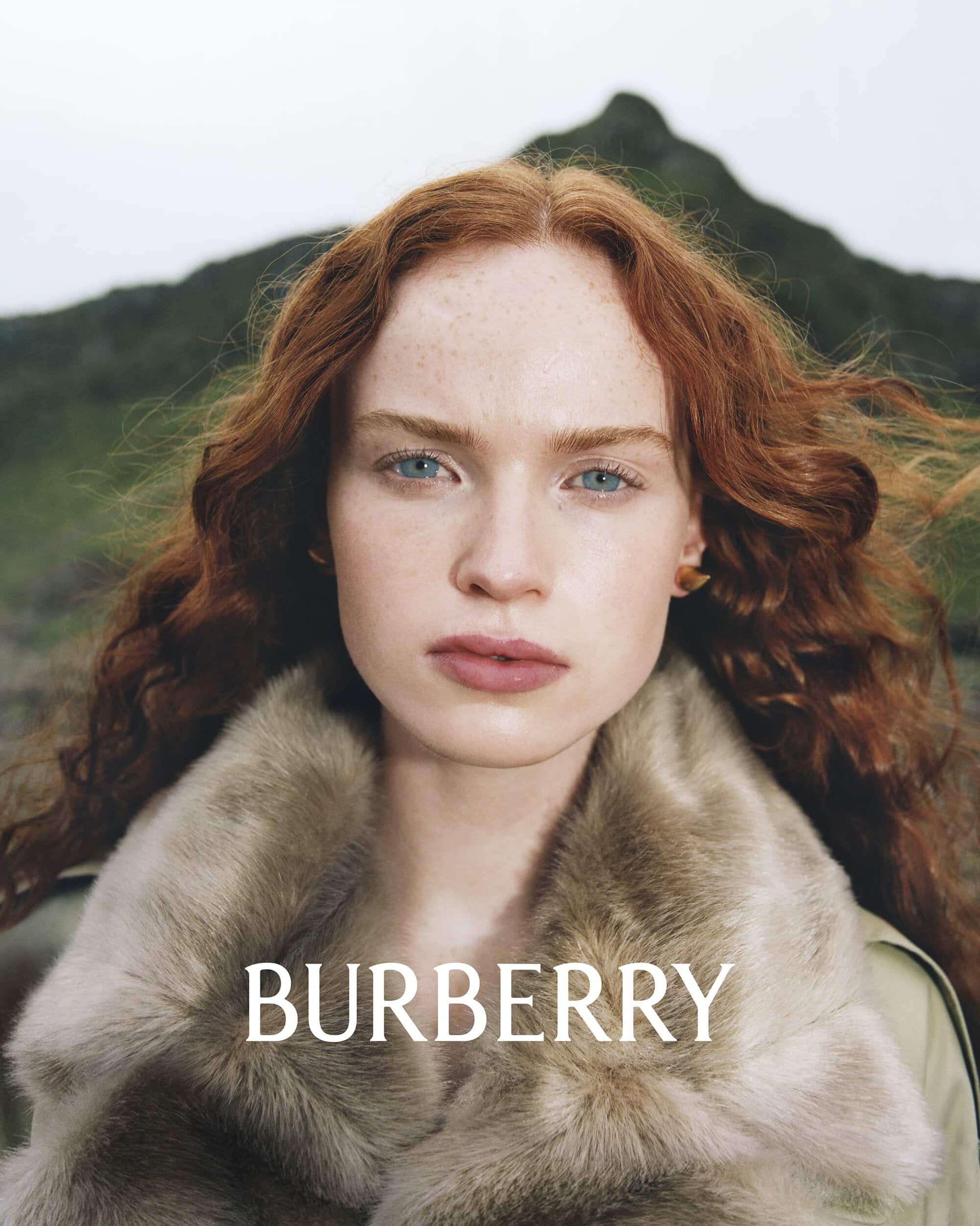 Portrait of a young woman with red hair and light eyes, wearing a fur coat against a blurred background of a green mountain, Burberry logo.