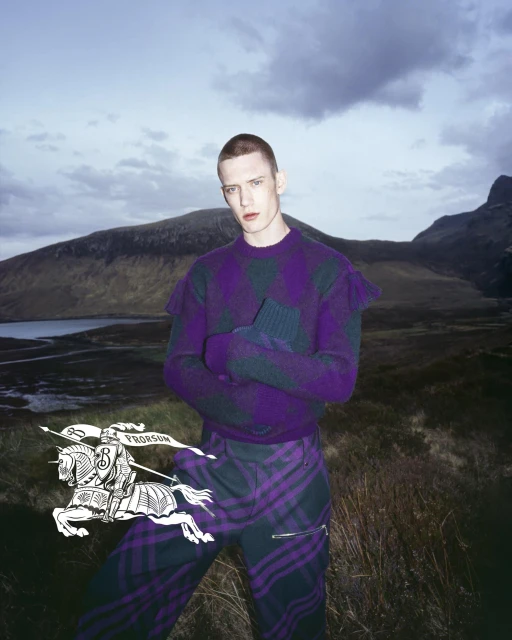 Man poses in purple plaid sweater and pants in outdoor mountain setting with a Burberry logo overlay, fashion forward and striking.