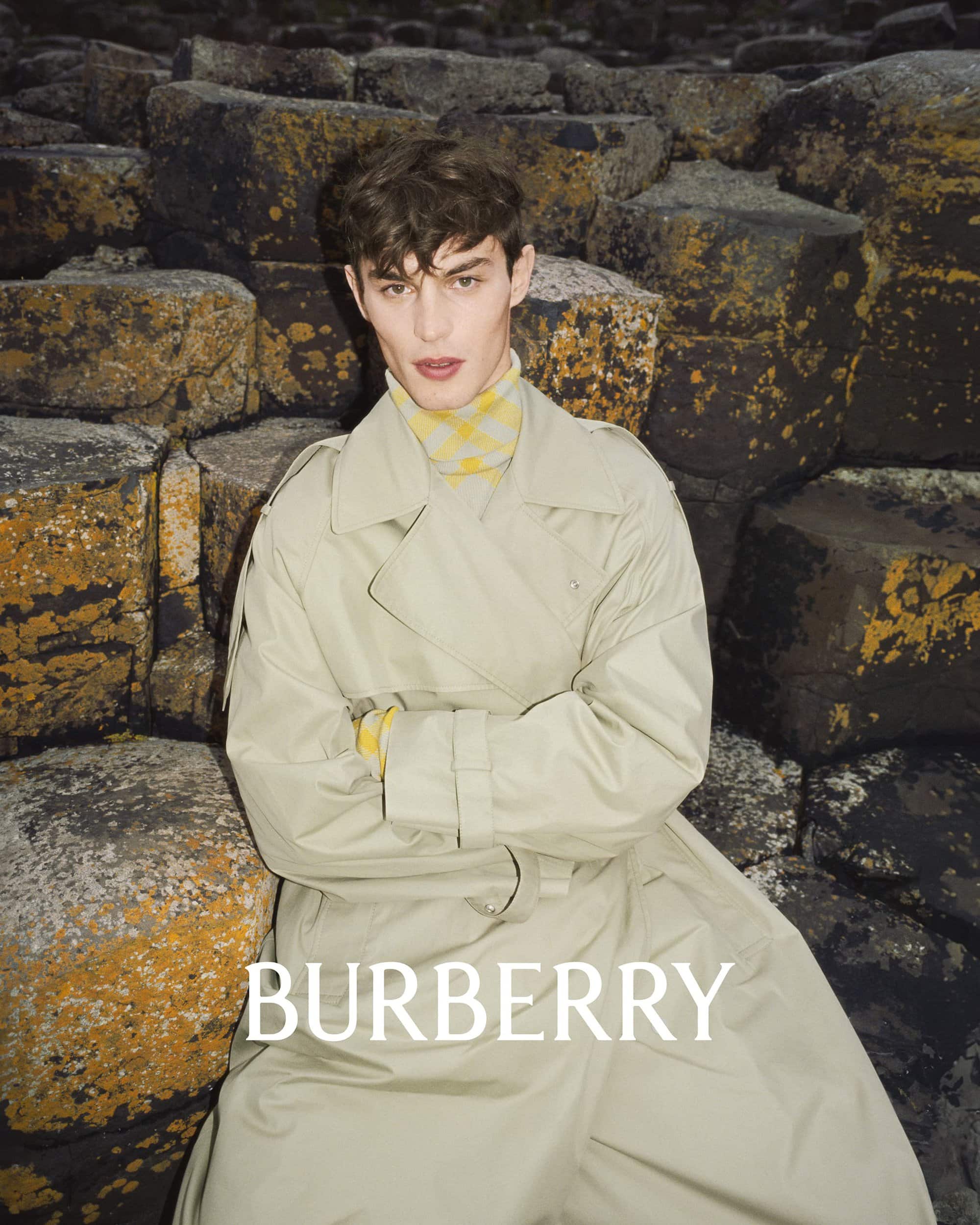A young man in a trench coat poses in front of a rock formation, showcasing a fashion editorial shot for the brand Burberry.