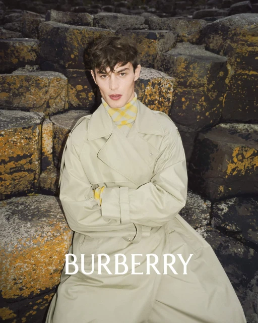 A young man in a trench coat poses in front of a rock formation, showcasing a fashion editorial shot for the brand Burberry.
