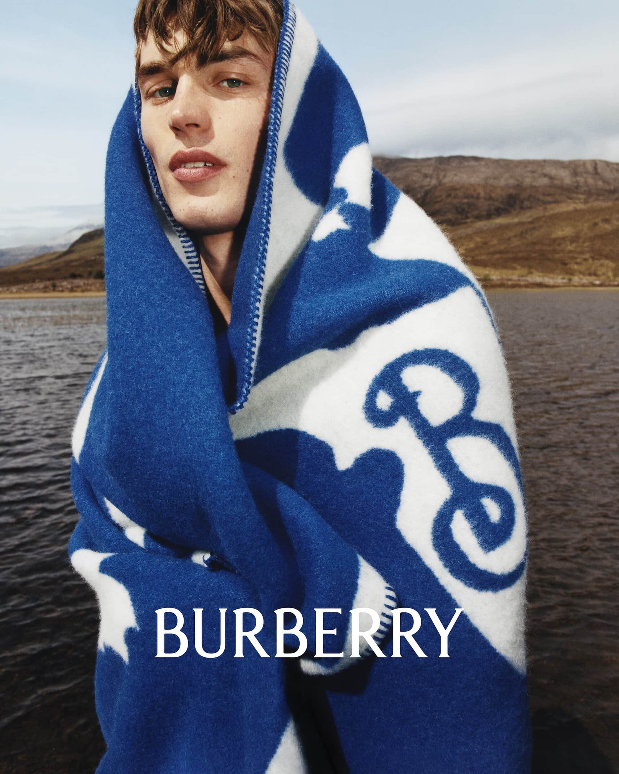 A blond male model is wrapped in a blue and white Burberry blanket with a logo near a lake, against a backdrop of hills and cloudy skies.