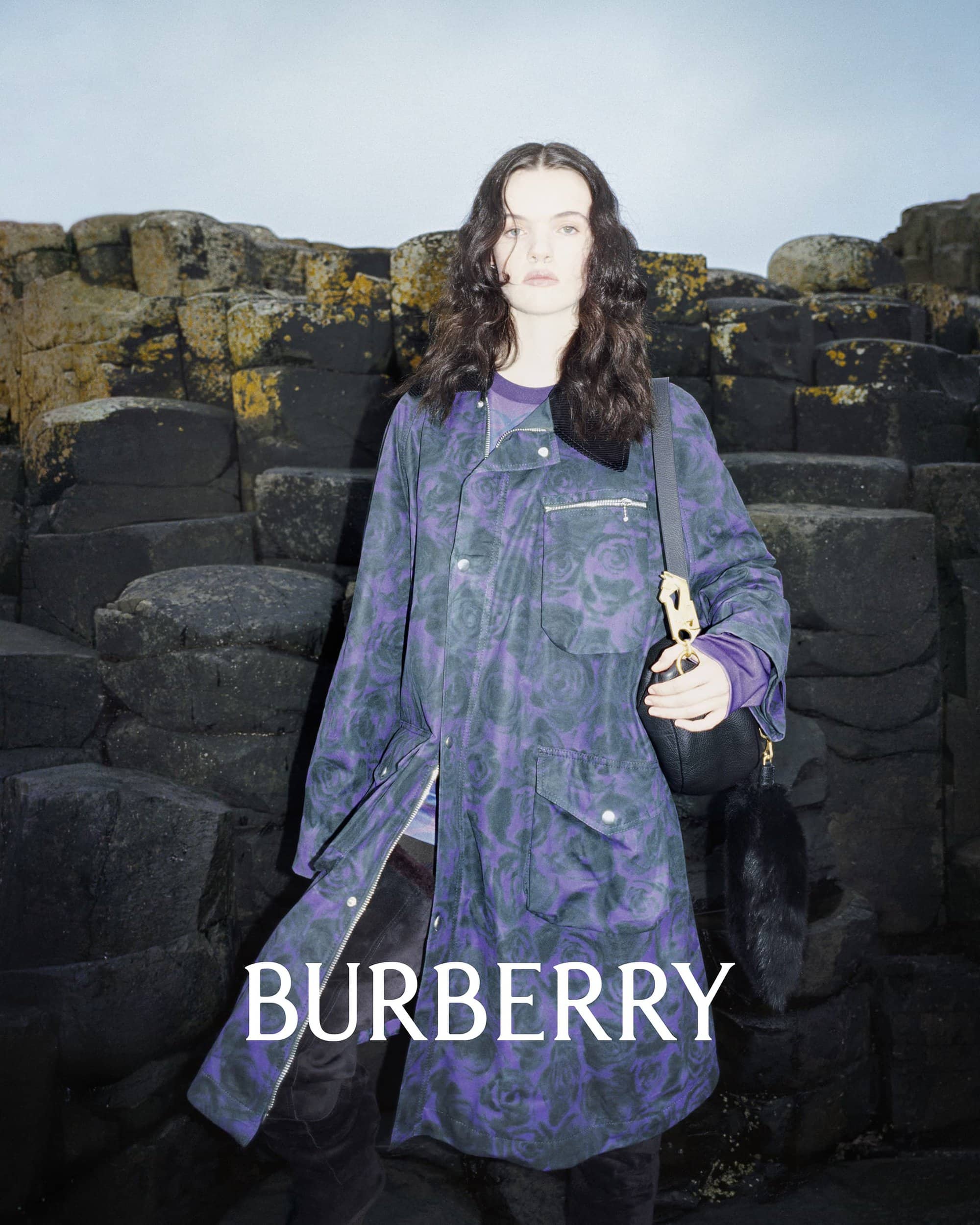A person with long brown hair in a purple jacket with a shoulder bag in front of a stacked rock formation, in a fashion editorial.