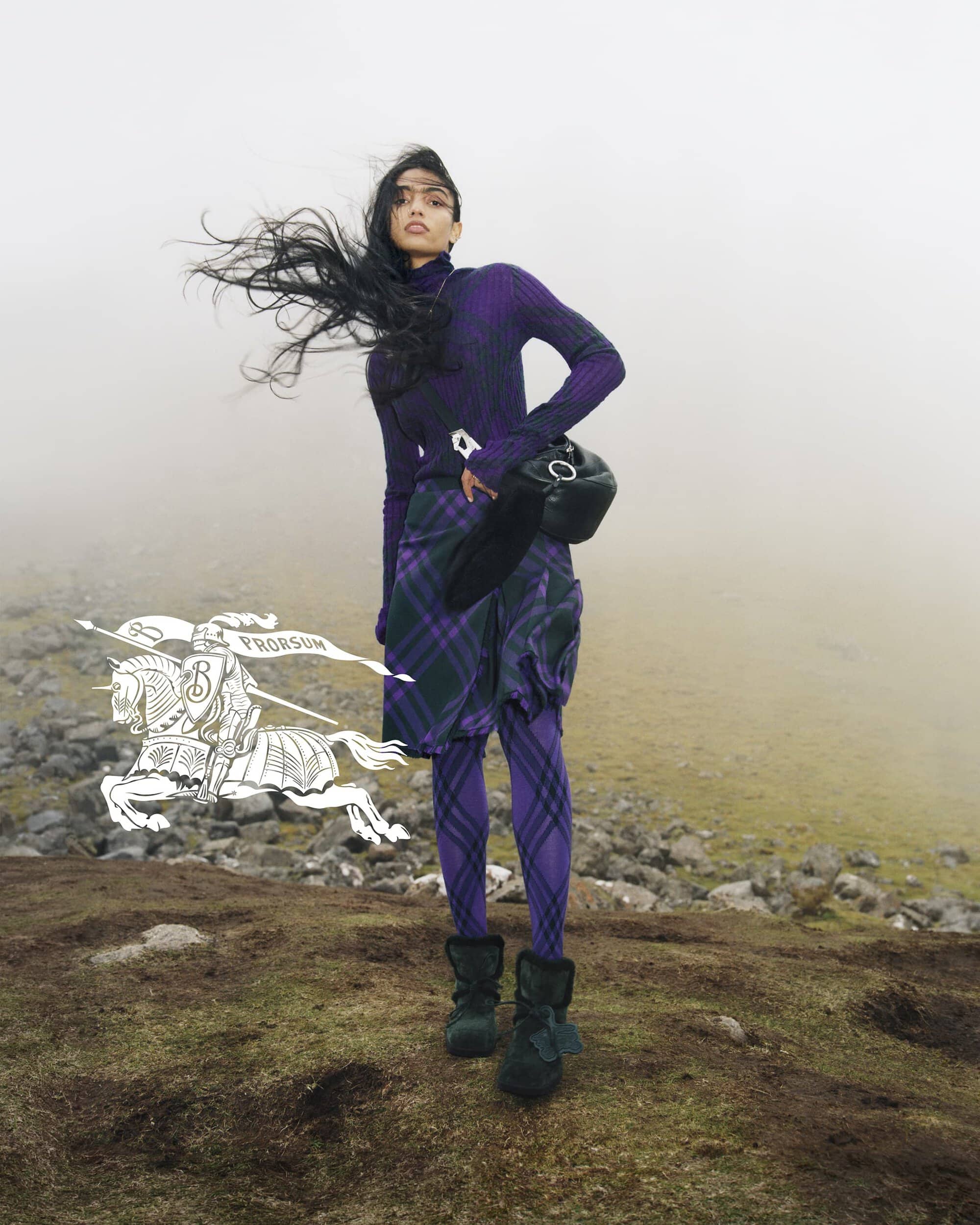 Fashion shot of a woman wearing a purple turtleneck, plaid skirt, and boots, standing outdoors with wind blowing her hair.