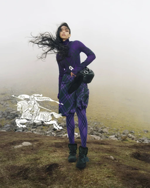 Fashion shot of a woman wearing a purple turtleneck, plaid skirt, and boots, standing outdoors with wind blowing her hair.