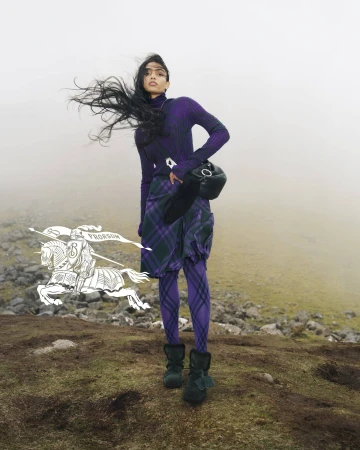 Fashion shot of a woman wearing a purple turtleneck, plaid skirt, and boots, standing outdoors with wind blowing her hair.