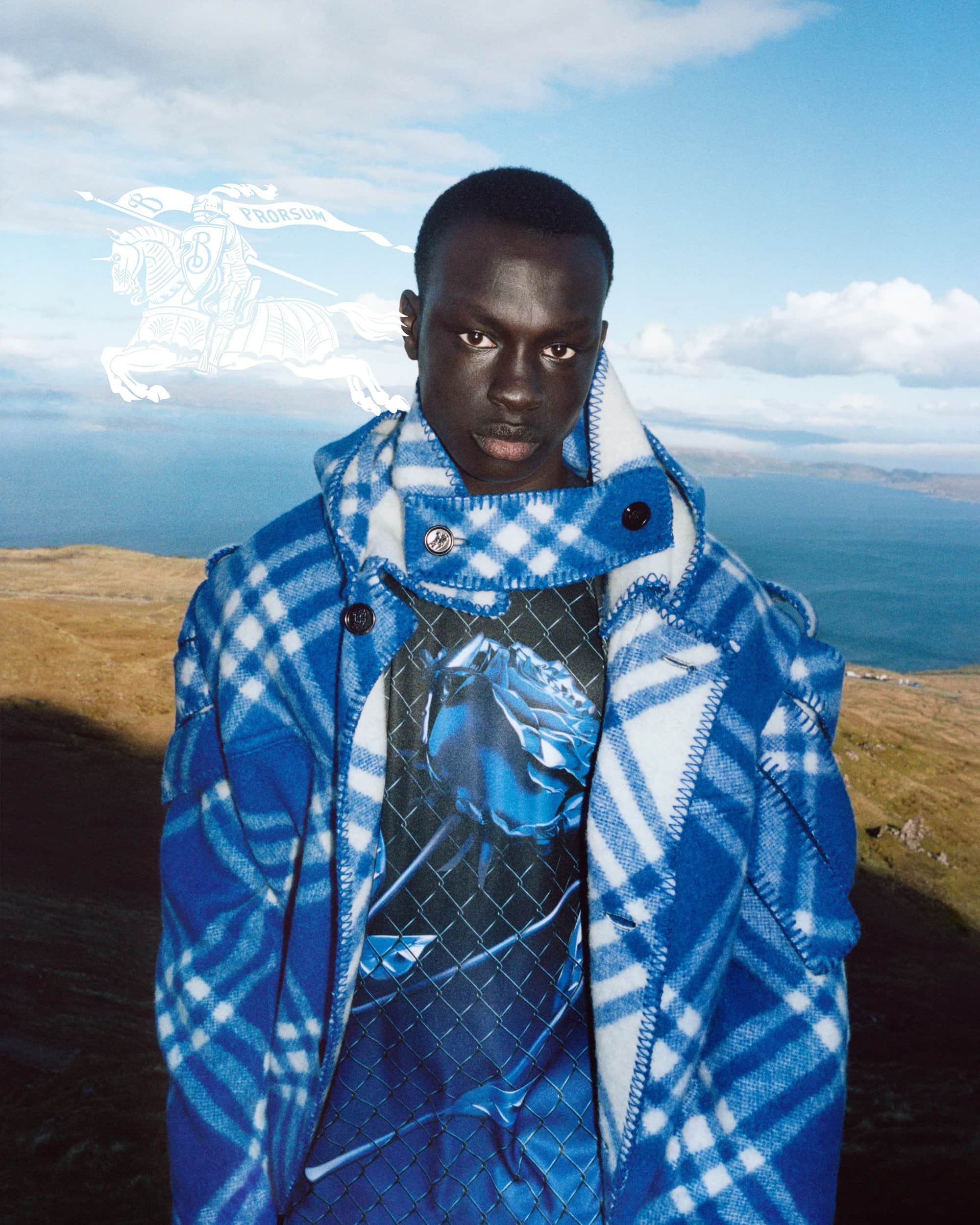 Fashion portrait of a Black male model wearing a plaid coat against a scenic mountain and sky background, with a vintage logo.