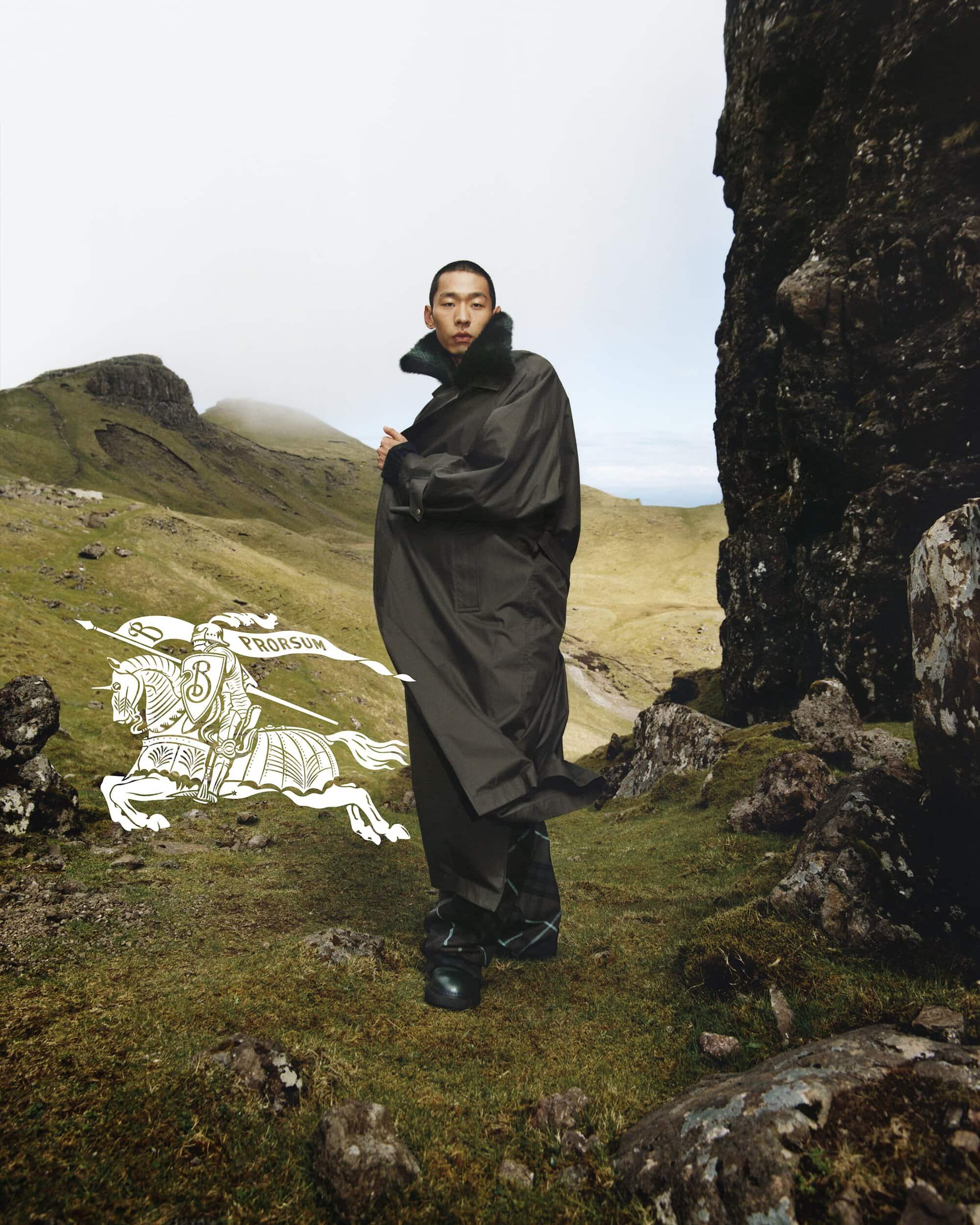 An adult male fashion model poses in a full-length coat against a backdrop of rocky terrain and grass hills in an outdoor fashion editorial.