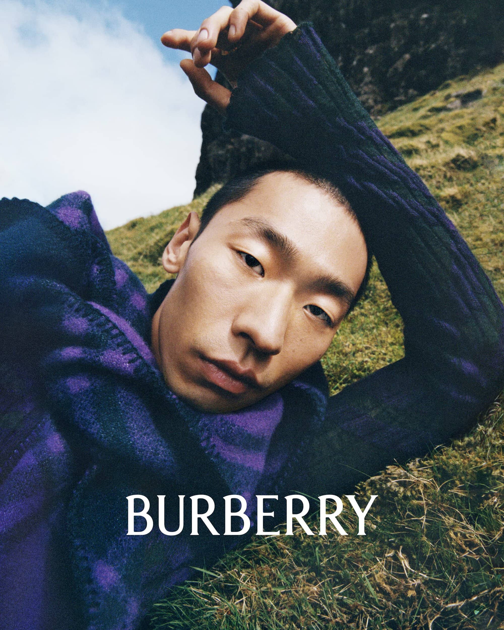 A stylish Asian male model poses in a Burberry sweater while lying on a grassy hillside, set against a backdrop of rocks and blue sky.
