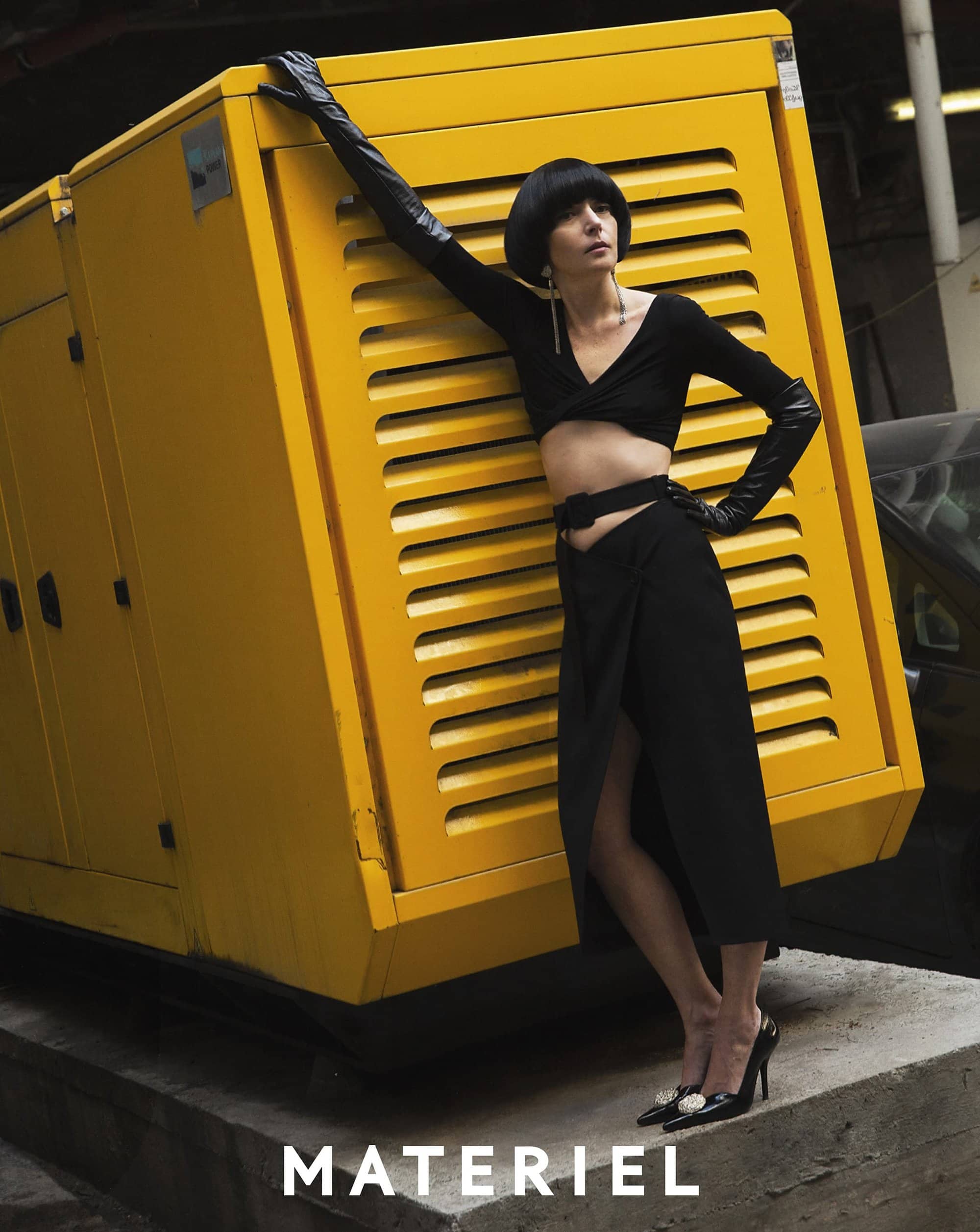 Fashion shot: woman in black crop top, skirt, gloves poses with yellow industrial generator in an urban outdoor setting, editorial style.