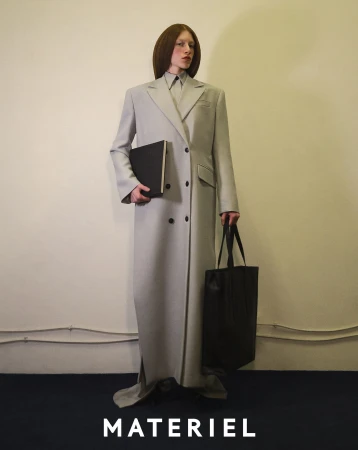 Model standing against a plain wall in a long grey coat holding a black portfolio and tote bag