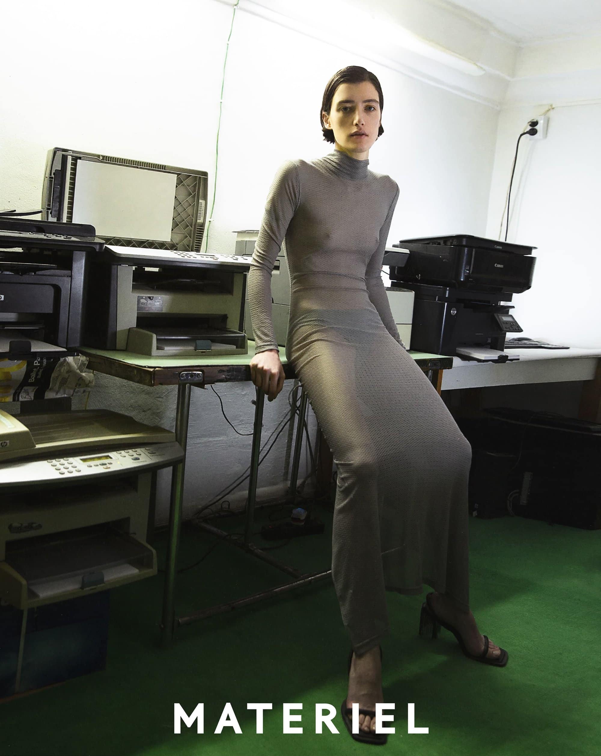 A woman in a grey turtleneck dress poses indoors in front of office equipment, looking directly at the camera with a neutral expression.