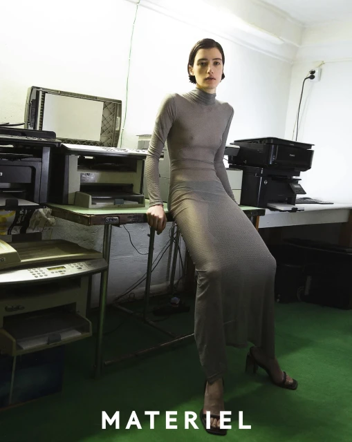 A woman in a grey turtleneck dress poses indoors in front of office equipment, looking directly at the camera with a neutral expression.