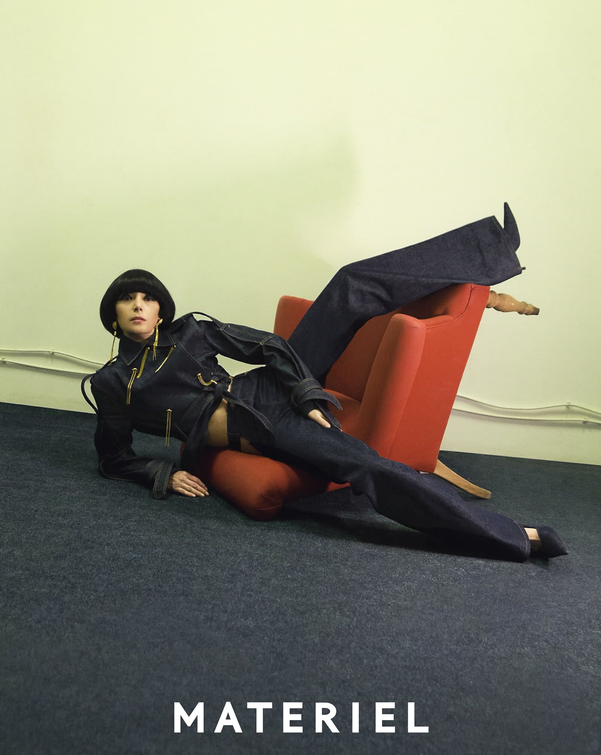 A woman in a dark denim outfit reclines on a red chair against a light green wall, showcasing a high-fashion editorial style.
