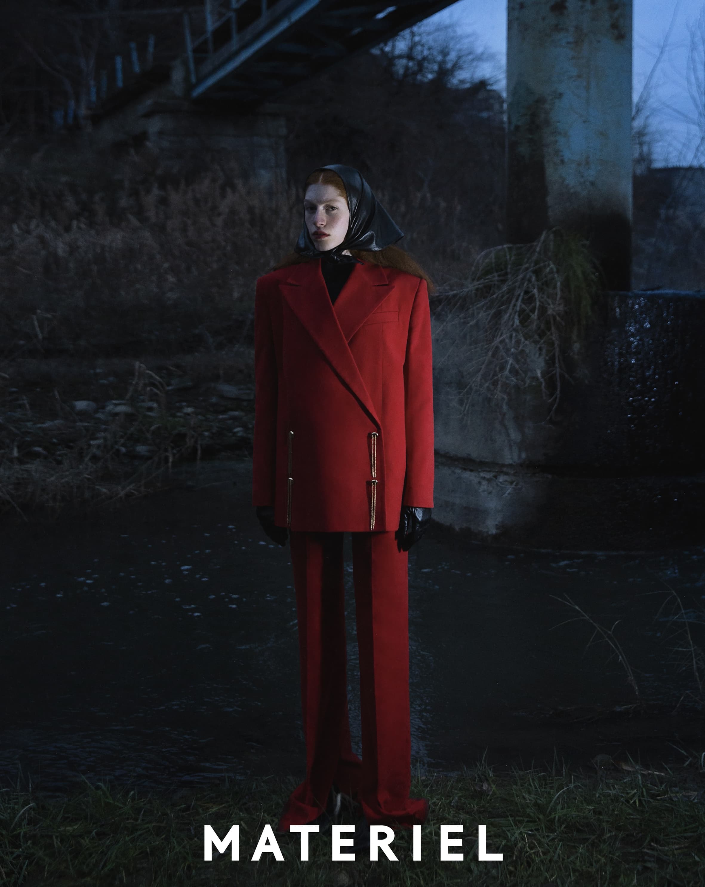 A full shot of a model in a red suit and headscarf standing in a dark outdoor environment next to a body of water, fashion shoot.
