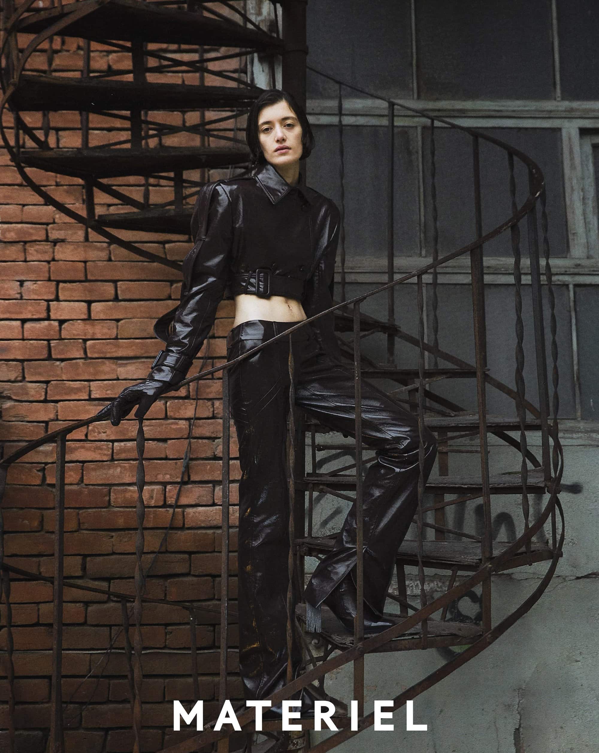 A woman poses on a spiral staircase wearing a black, shiny outfit of a cropped jacket, pants, and gloves, with a brick wall backdrop.