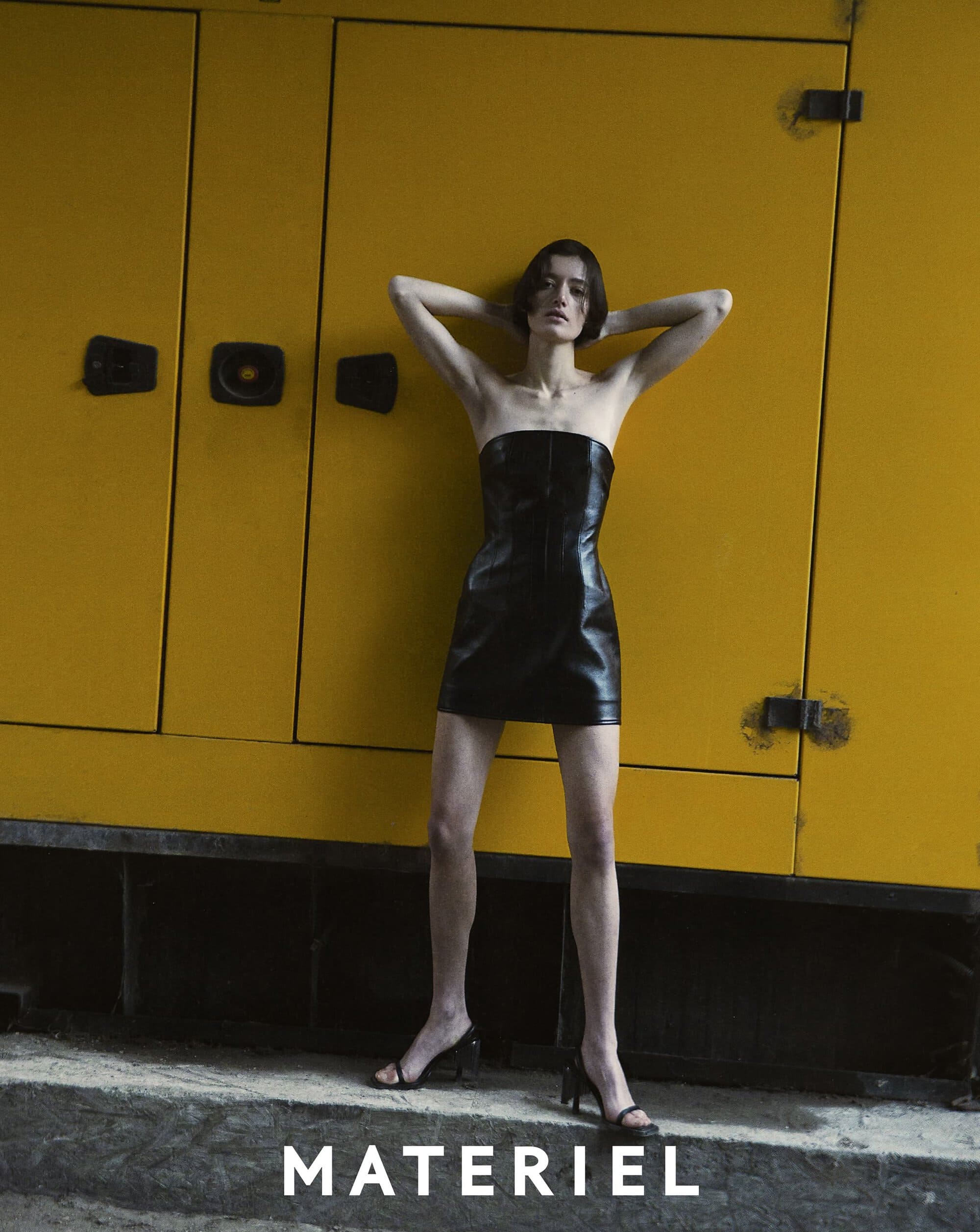 Fashion photo of a slender woman in a black mini dress, standing against a yellow industrial generator. Studio lighting, editorial style.
