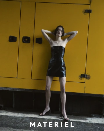 Fashion photo of a slender woman in a black mini dress, standing against a yellow industrial generator. Studio lighting, editorial style.