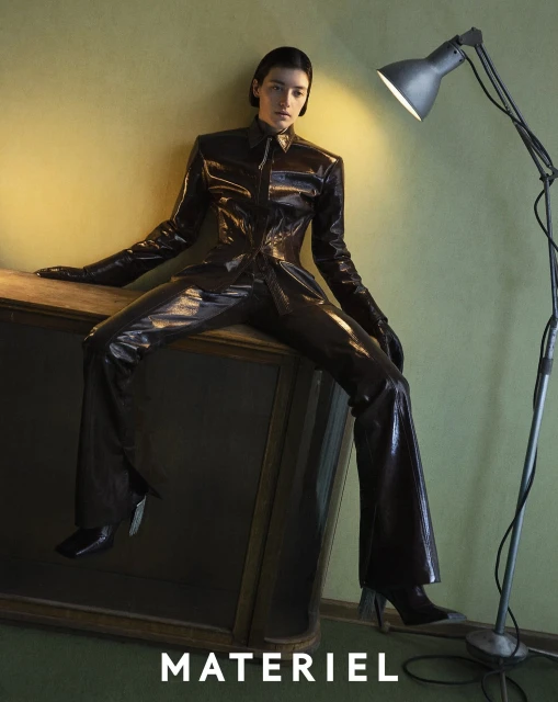 A fashion model poses in a shiny black pantsuit on a wooden cabinet against a yellow-green wall, illuminated by a lamp, showcasing chic style.
