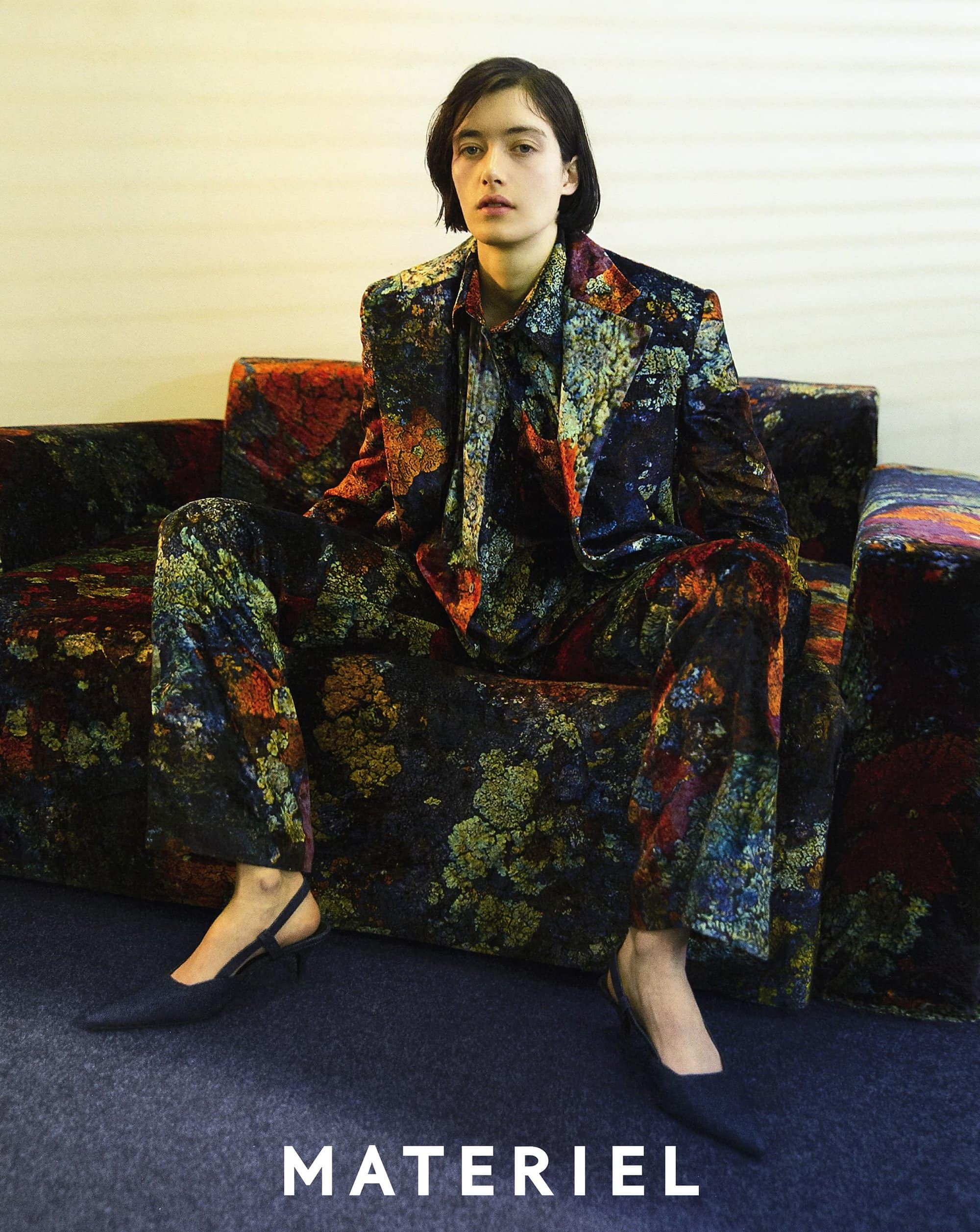 A woman poses on a floral couch in a floral suit, wearing black heels, sitting indoors, in a medium studio shot with neutral lighting.