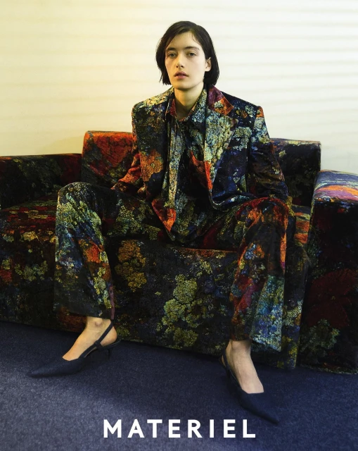 A woman poses on a floral couch in a floral suit, wearing black heels, sitting indoors, in a medium studio shot with neutral lighting.