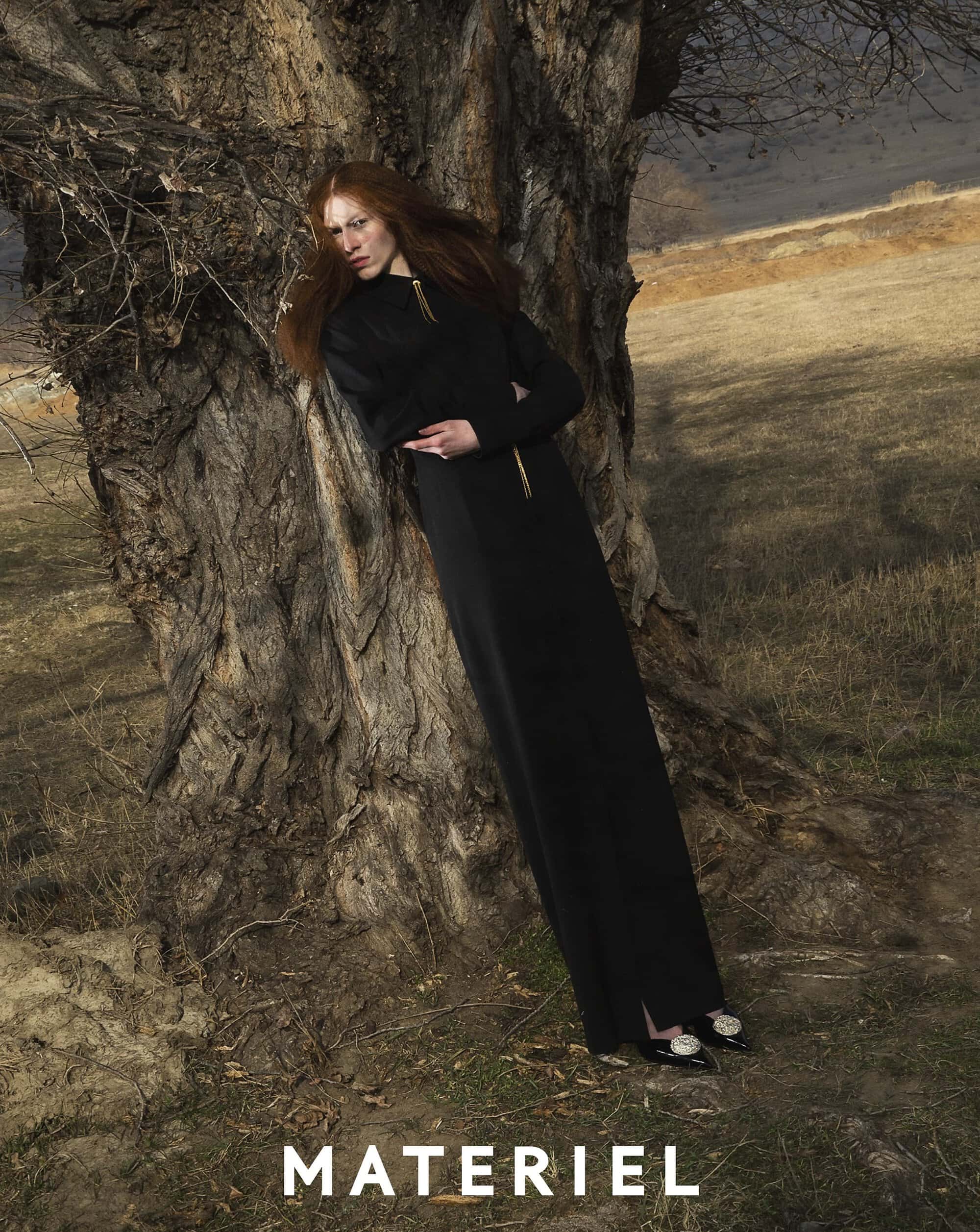 A redhead woman in a long black dress leans against a gnarled tree in a field, creating an editorial outdoor fashion portrait.