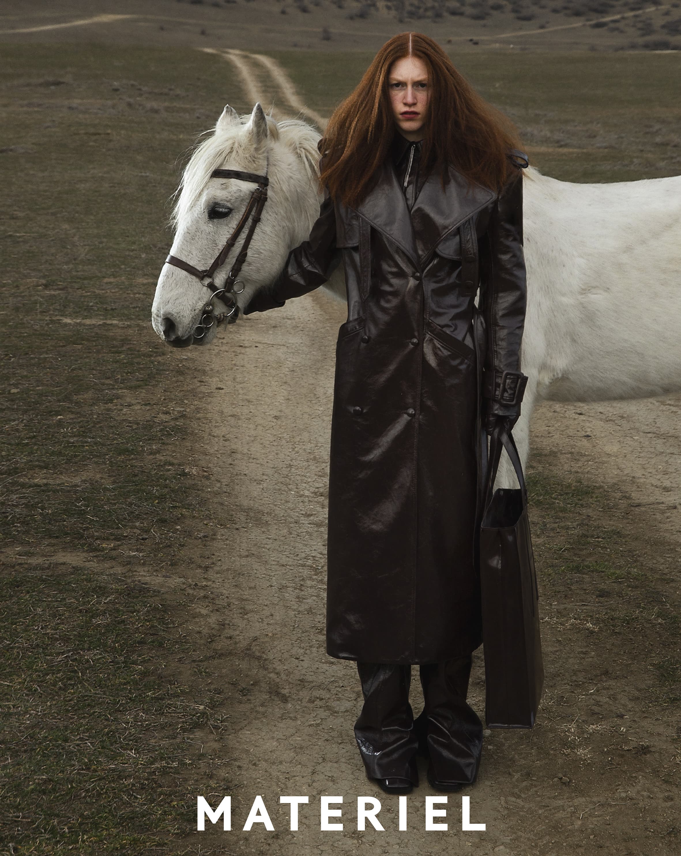 A person in a black coat stands in a field with a white horse on a path, creating a striking and fashionable visual narrative.