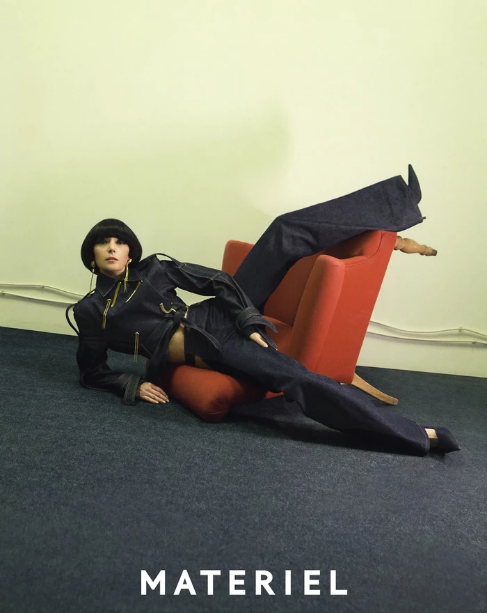 Model in dark denim jacket and matching trousers reclining against a red armchair on a dark carpet for Materiel Pre-Fall 2023 campaign