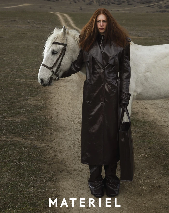Model wearing a dark long leather coat standing next to a white horse on a dirt path in an open field landscape