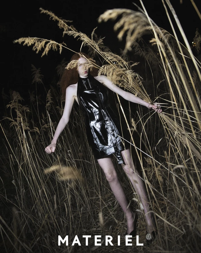 Model in black latex dress holding tall dried reeds outdoors at night for Materiel