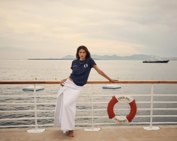 A woman in a navy T-shirt and white wrap skirt poses on a cruise ship with the ocean, a yacht, and the coastline in the background.