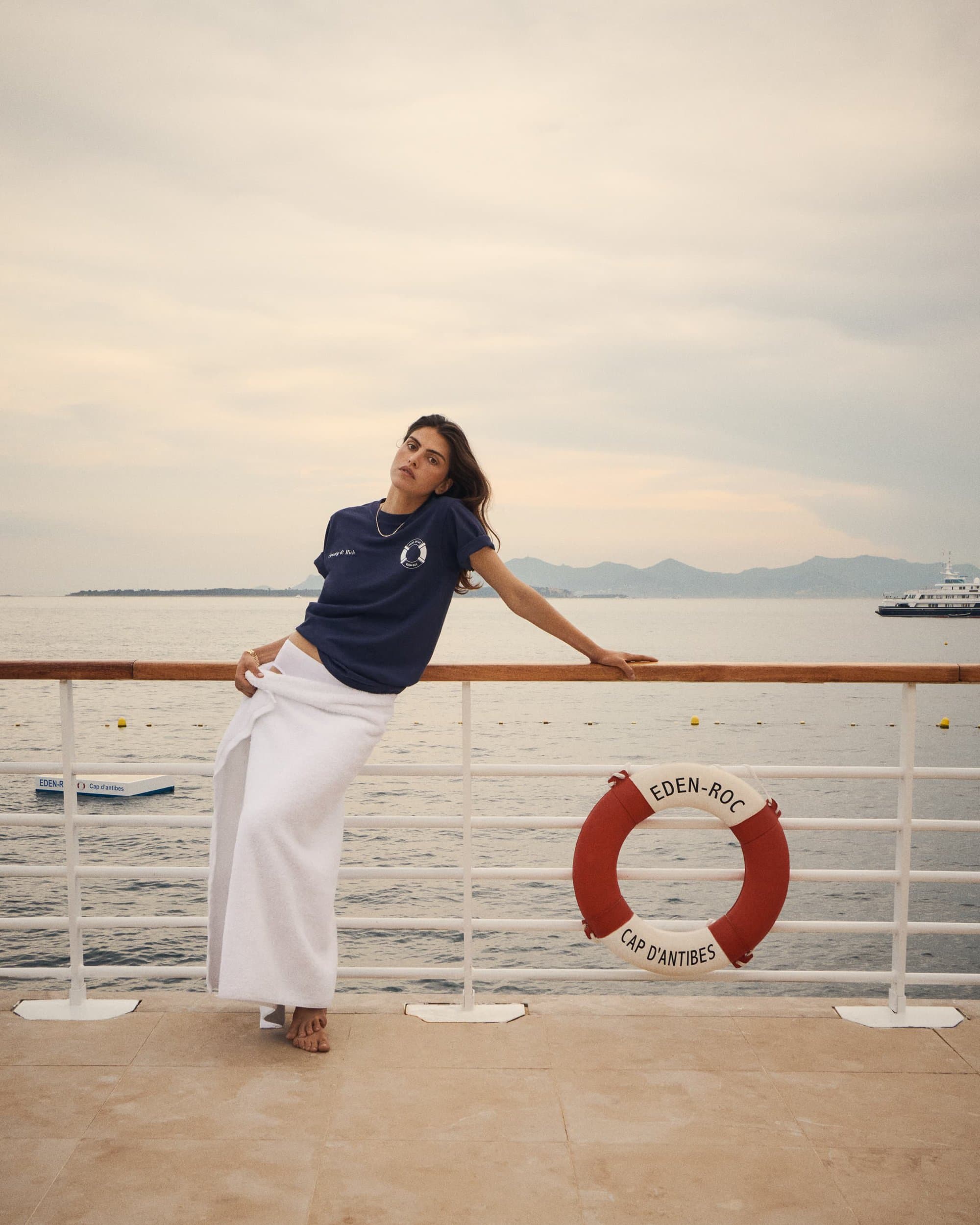 A woman in a T-shirt and wrap skirt leans on a ship's railing overlooking the ocean and distant mountains under a cloudy sky.