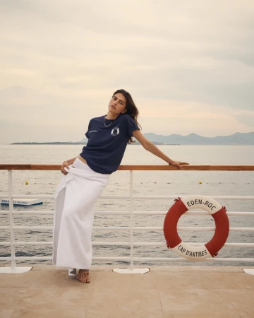 A woman poses on a boat deck wearing a navy t-shirt and white skirt with the ocean, mountains, and sky visible in the background.