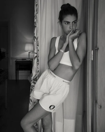 Black and white portrait of a woman in a sporty top and boxer shorts leaning against a doorframe with a serious facial expression.