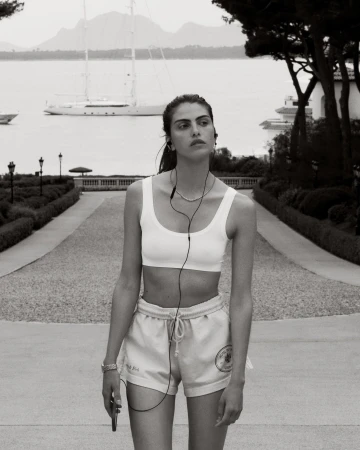 Woman in athletic wear walks on a stone path with a large sailboat visible in the background. She's wearing headphones and holding a phone.