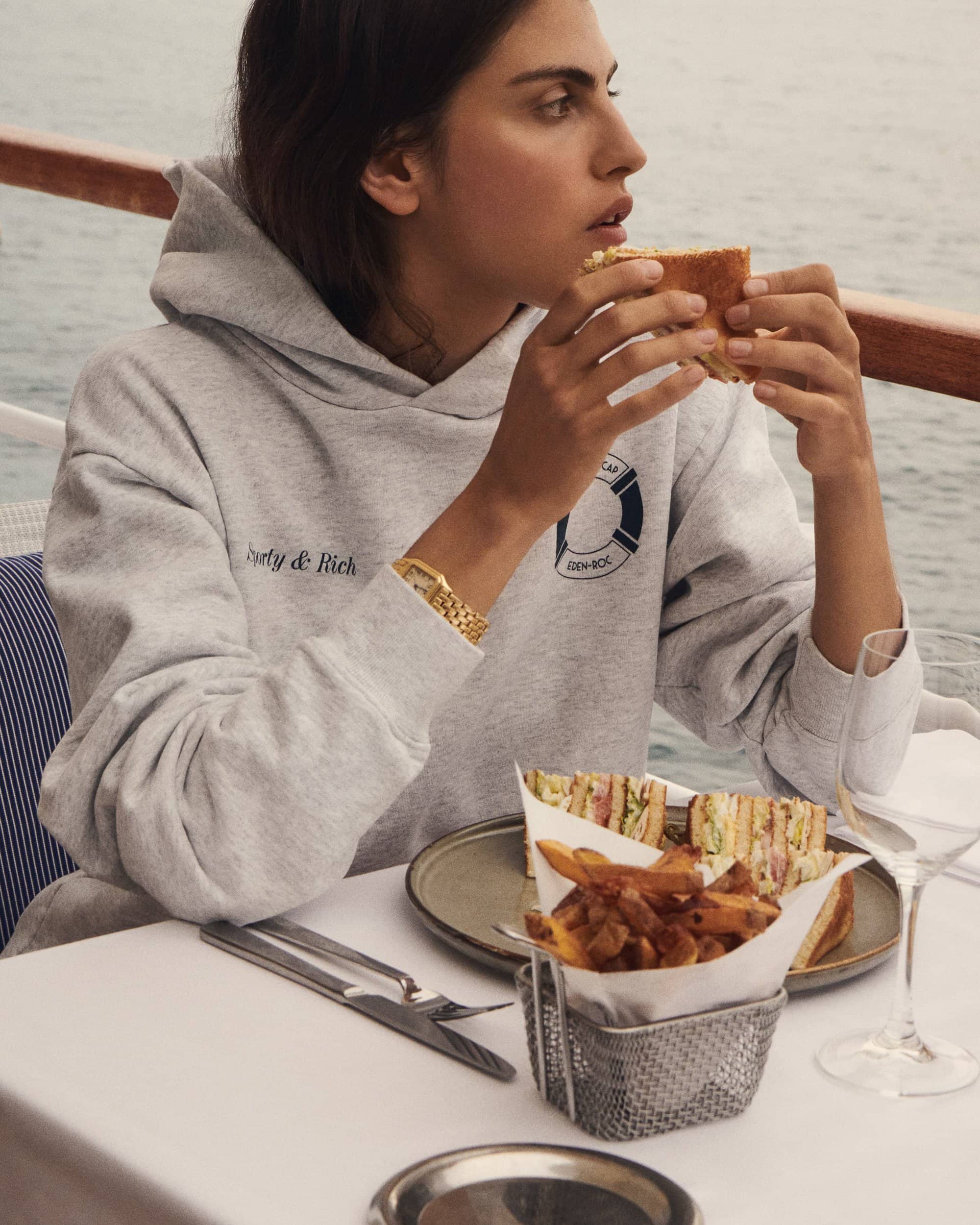 A woman in a grey hoodie sits at a table on a boat, eating a sandwich with fries and ocean in background in a lifestyle shot.
