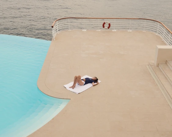 Woman in swimwear relaxes on a towel by a pool overlooking the ocean; poolside setting with turquoise water and neutral colors.