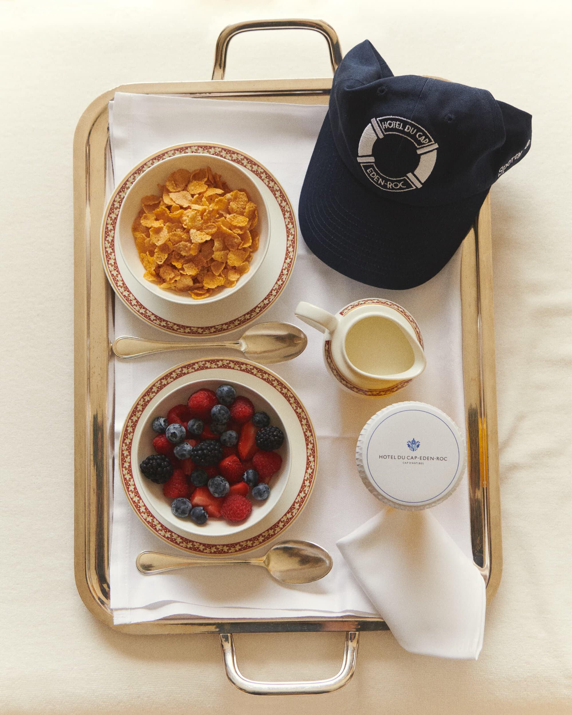 Overhead shot of a silver tray with breakfast items: cereal, berries, milk, and a baseball cap, all on a white linen.