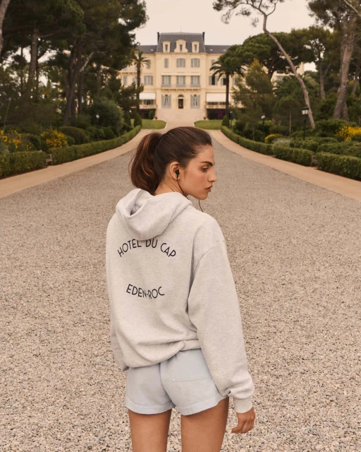 A woman stands on a path leading to a grand hotel, wearing a hoodie with 'Hotel du Cap Eden Roc' printed on the back.