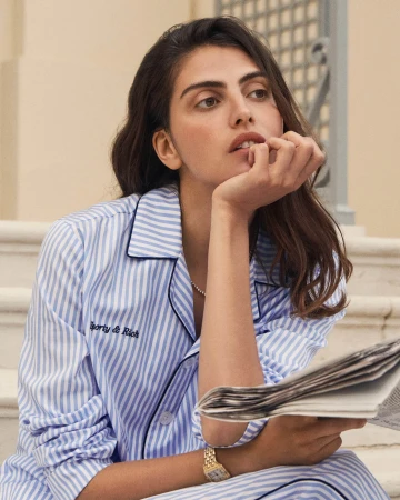 Brunette woman in striped pajamas sits on steps outdoors reading newspaper, accessorized with gold watch and pearl necklace.