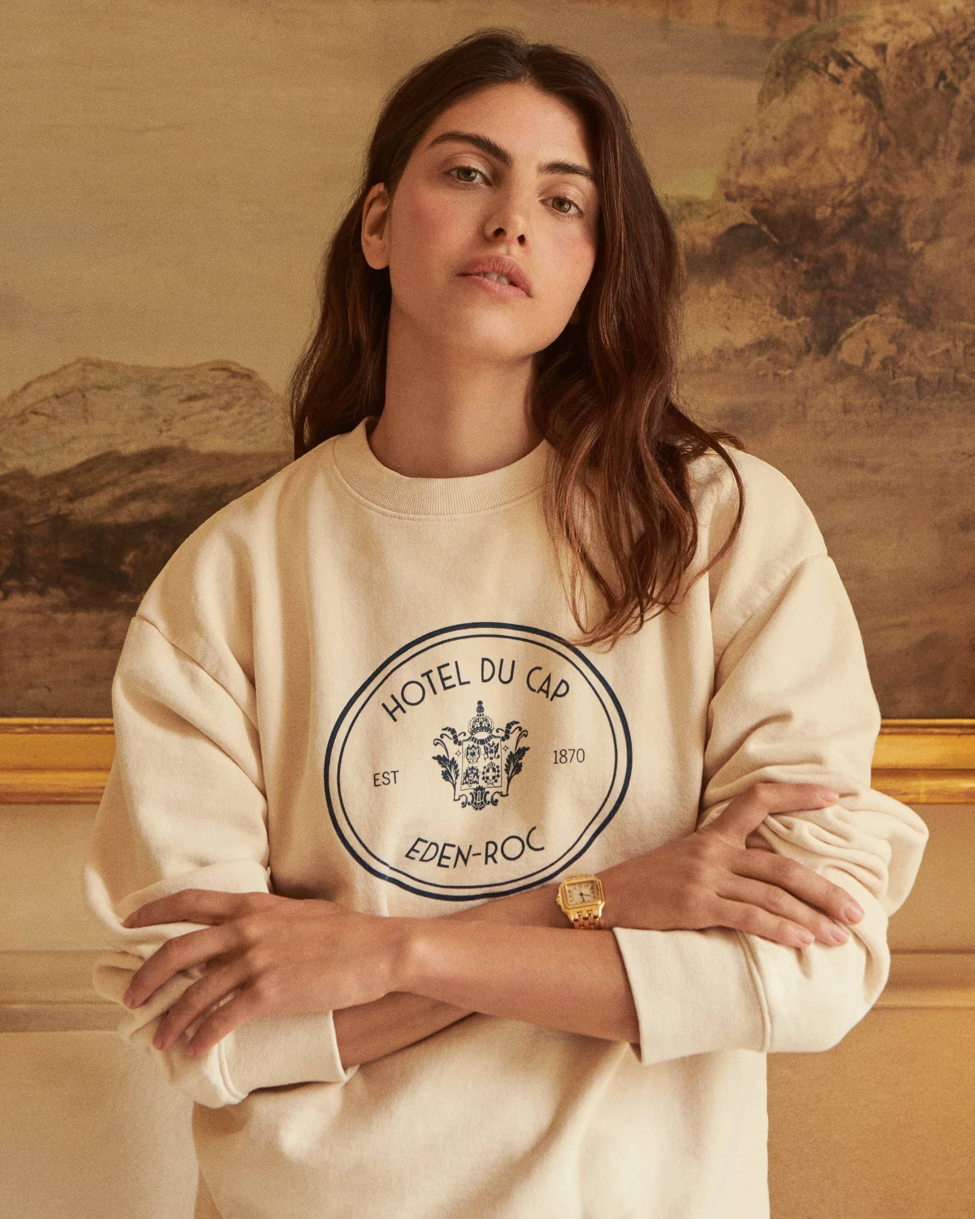 Brunette woman wearing a cream Hotel Du Cap sweatshirt with arms crossed, standing indoors against a muted painted wall.