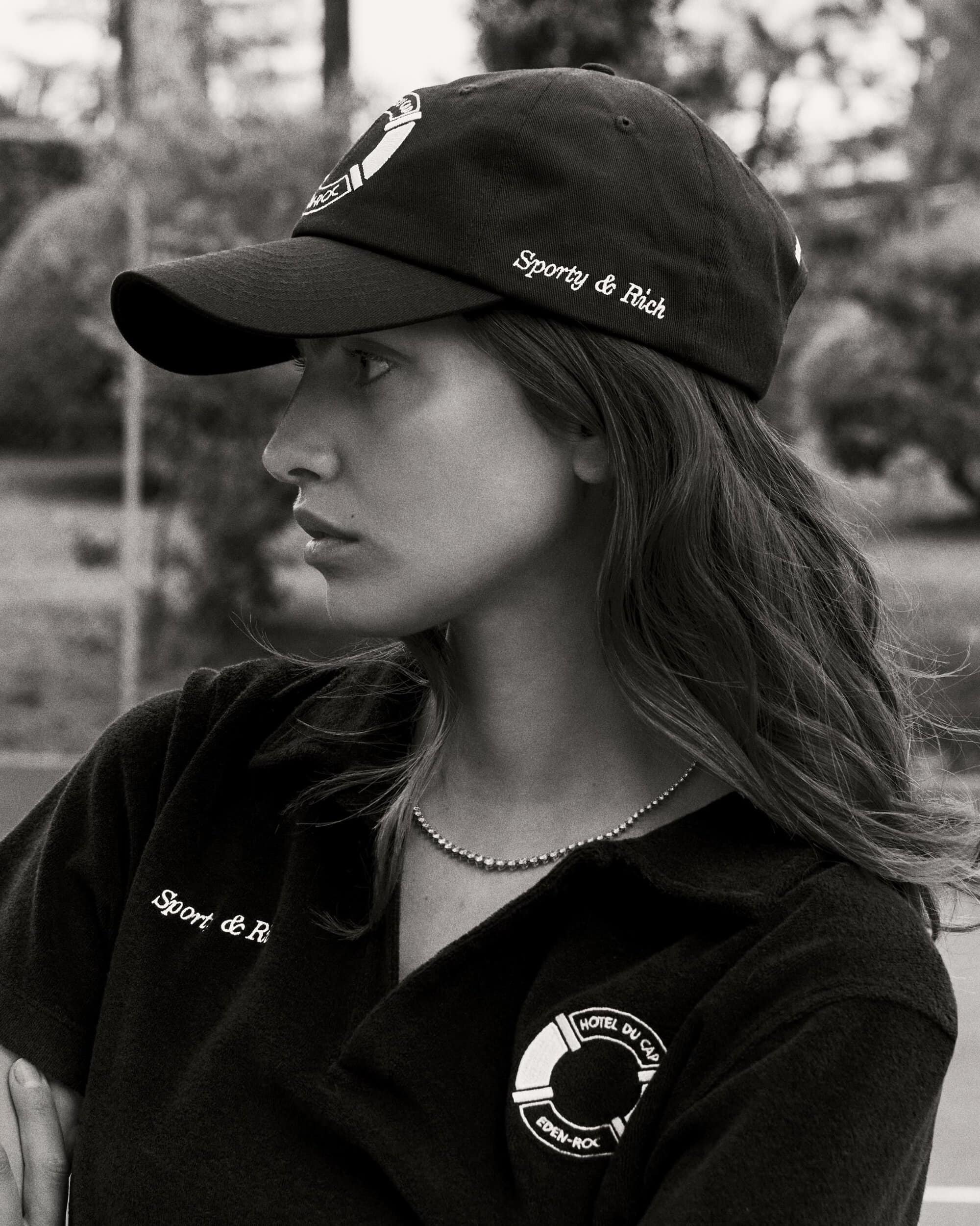 Woman in 'Sporty & Rich' baseball cap and polo shirt with 'Hotel du Cap-Eden-Roc' logo in a monochrome outdoor setting.