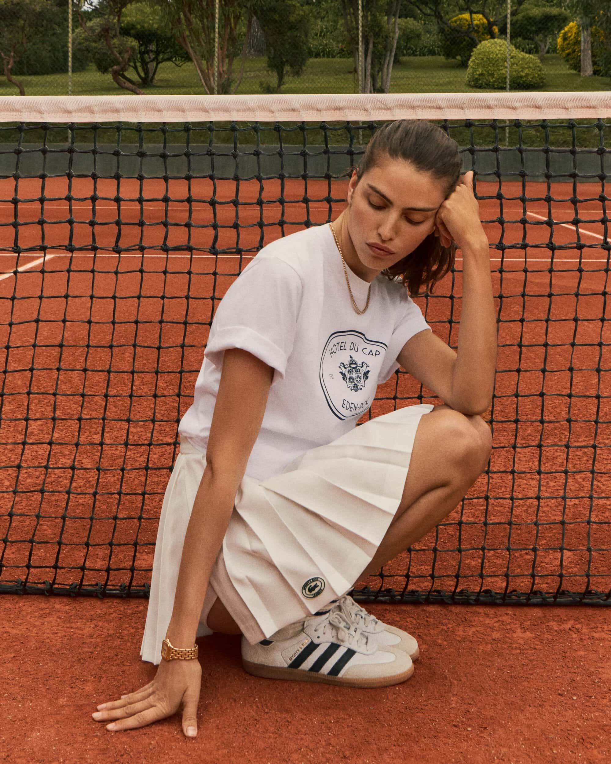 Brunette woman poses on a clay tennis court, wearing a white t-shirt, pleated skirt, and athletic shoes, with a net in the background.