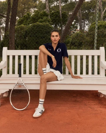 Brunette woman in tennis outfit sits on white bench with racket at outdoor court with green trees, wearing navy polo and white skirt.