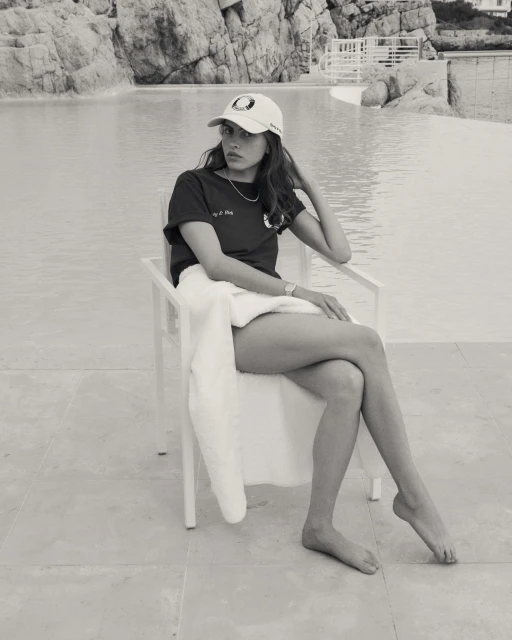 Black and white image of a young woman in a baseball cap sitting on a chair next to a pool in an outdoor setting.